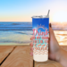 Close-up of woman holding tumbler with beach in background and sunny, relaxed vibes.